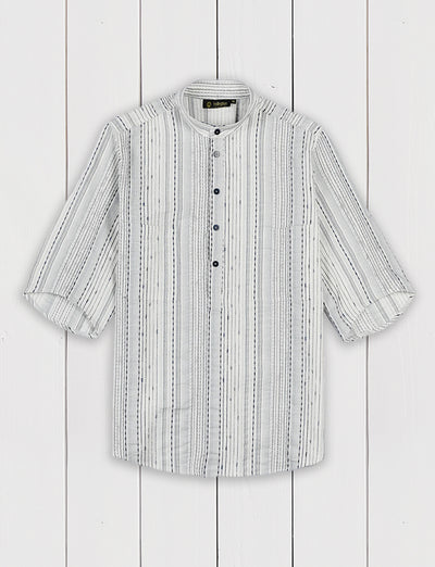 Cette chemise est coupée dans une toile Ulysse Pila, maison spécialisée dans les soieries orientales fondée à Lyon en 1867. De type « ethnique », elle est profondément texturée. Ses rayures verticales sont formées par une variation de fils écru, noisette et gris fumée qui donnent l’impression d’une broderie. L’intensité des couleurs et des reliefs obtenus dépend de la densité en fils et de la forme de chaque ligne.